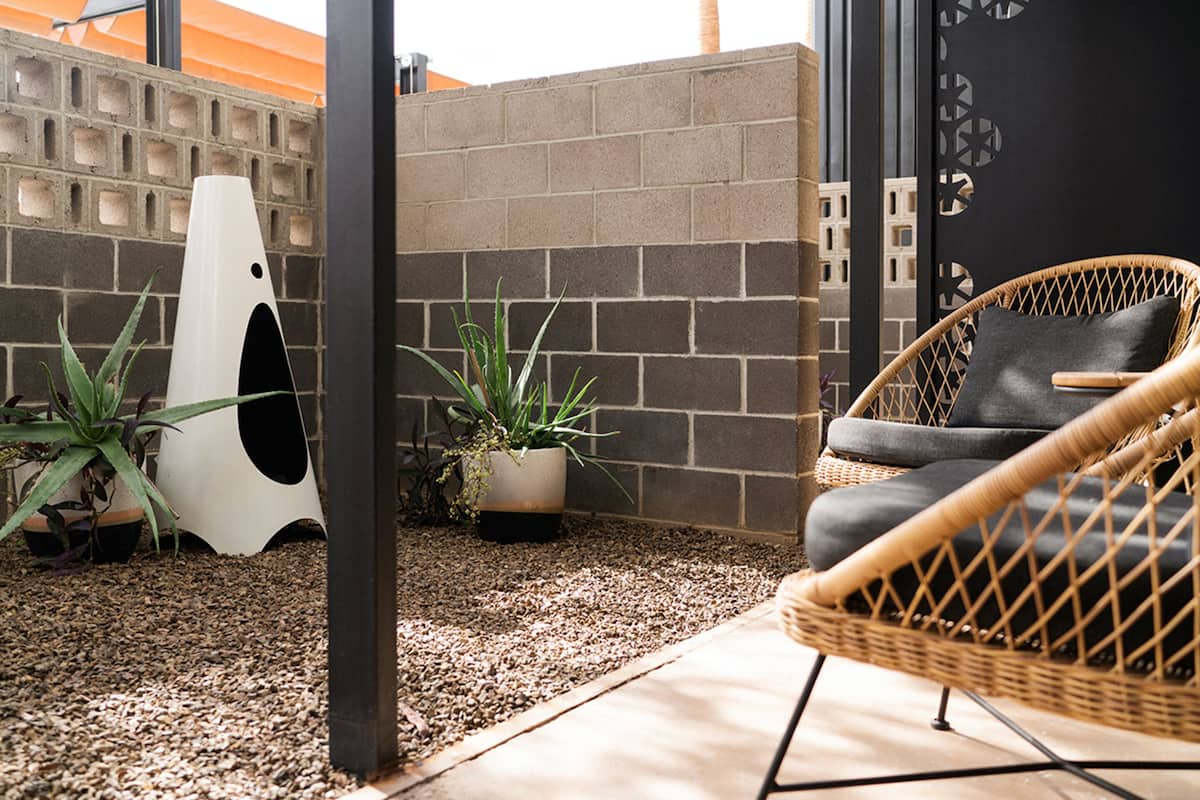 A wicker chair with a cushion, a potted plant, and a white birdhouse against a brick wall.
