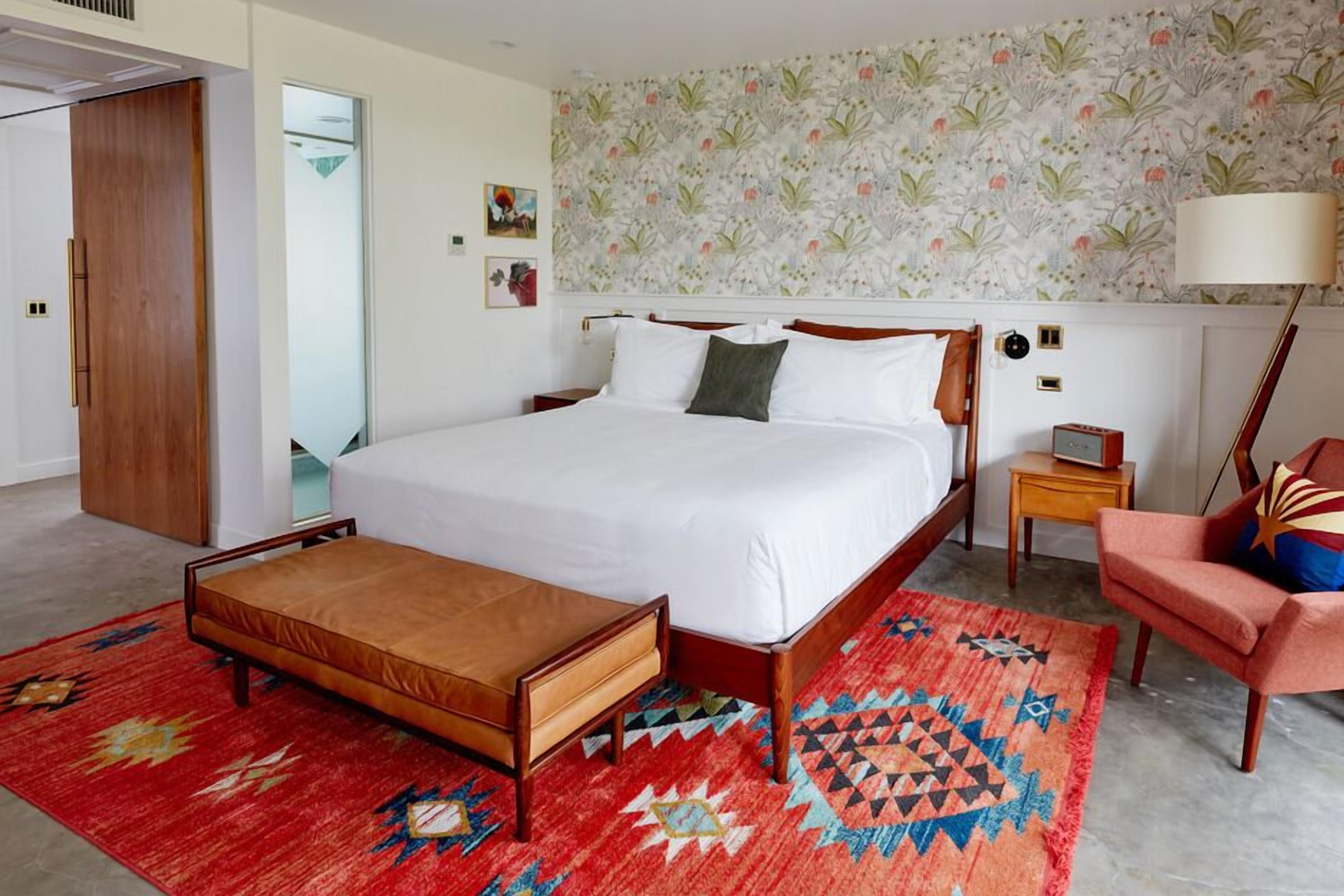 A modern bedroom with a large bed, a wooden bench, a patterned rug, and a chair.