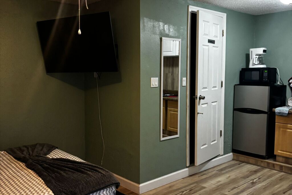 A compact room with a bed, a television mounted on the wall, a door leading to a kitchen area with a refrigerator and microwave.