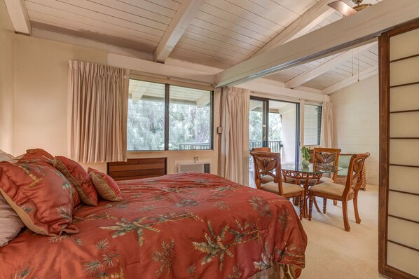 A bedroom with a large bed, a dining table with chairs, and a balcony with a view of trees.