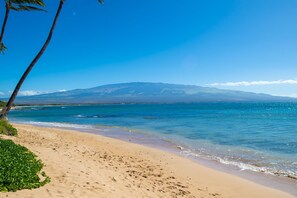 A beach with clear blue water, a sandy shore, and a mountain in the background.