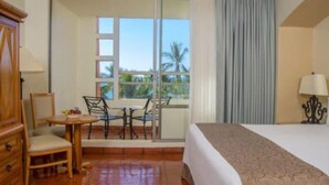 Une chambre d’hôtel comprenant un lit, un bureau avec des chaises, une fenêtre donnant sur des palmiers et une armoire en bois.