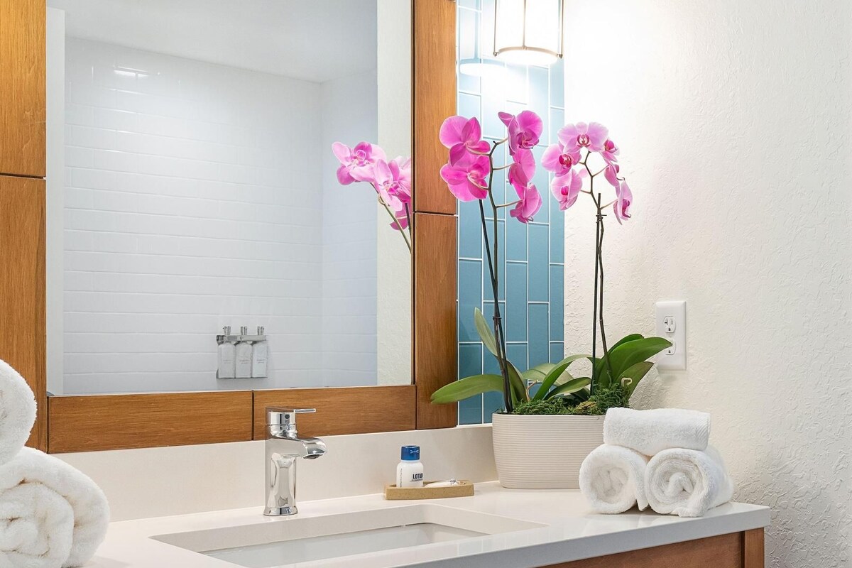 A bathroom with a large mirror, white sink, wooden vanity, and a potted orchid plant.