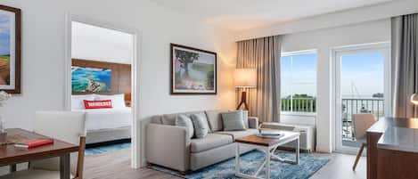 A modern hotel room with a sofa, coffee table, and a dining area. There is a large window with a view of the outdoors.
