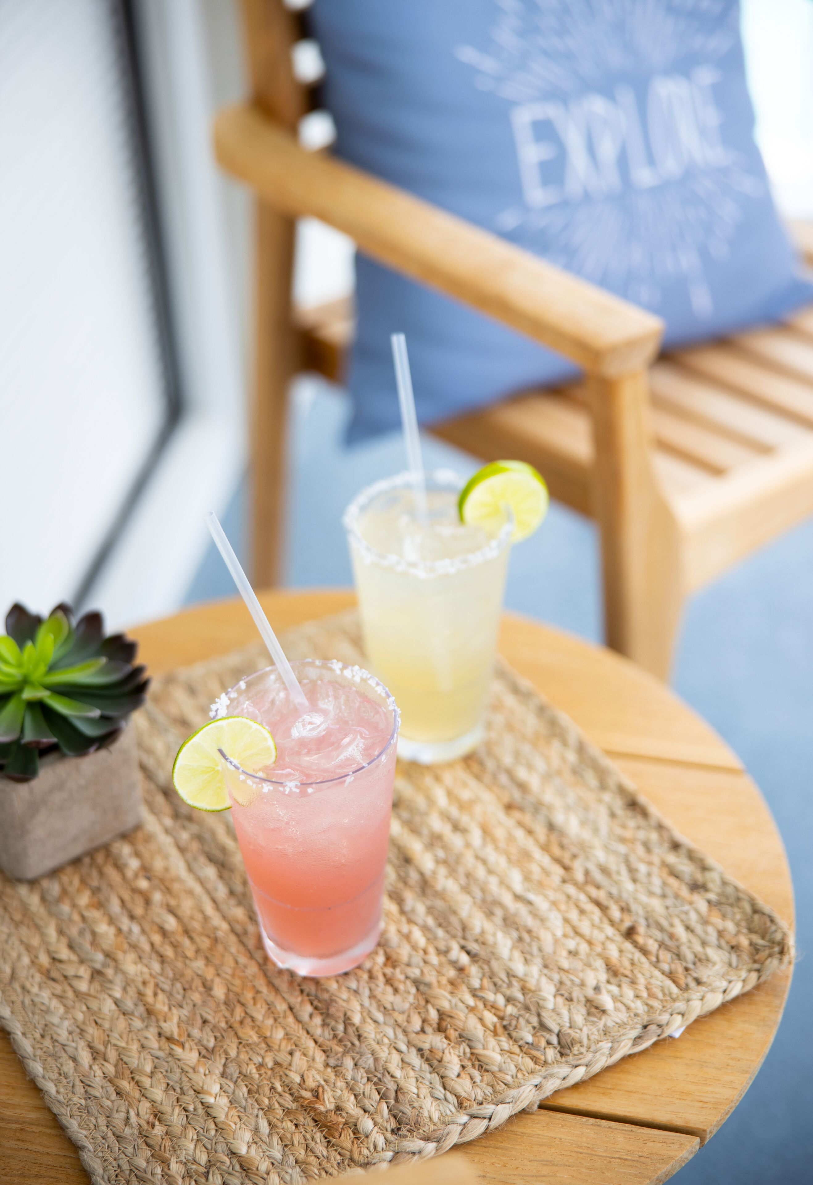 Two drinks on a woven placemat with a chair and a cushion in the background.