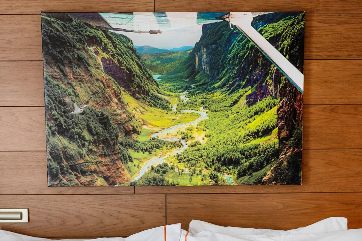 Een hotelkamer met een houten hoofdbord, een groot landschap van een vallei en een rood kussen met de tekst 'WANDERER'.