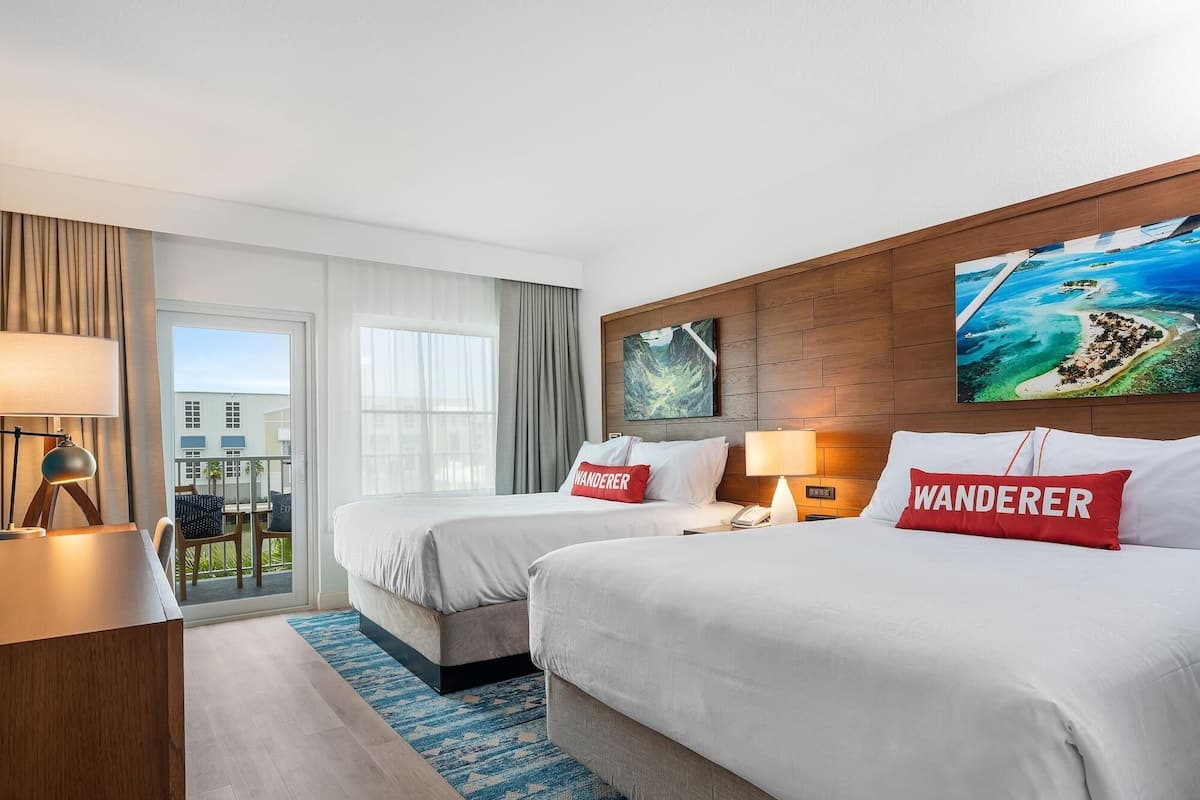 A hotel room with two beds, a desk, a chair, a window with a view, and a painting on the wall.