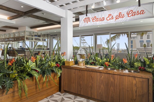 A reception area with a wooden counter, tropical plants, and a sign reading "Mi Casa Es Su Casa". - Compass Hotel by Margaritaville Anna Maria Sound (Bradenton)