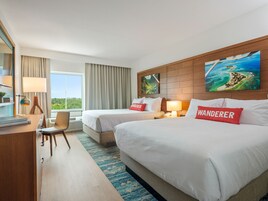 A hotel room with two beds, a desk with a lamp, a chair, and a large window with curtains.