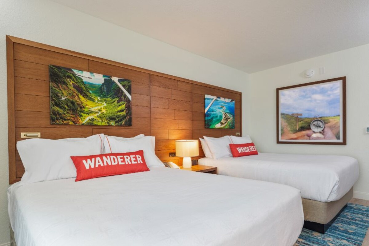 A hotel room with two beds, each with a pillow labeled 'WANDERER,' a wooden headboard, and a wall-mounted painting of a landscape.