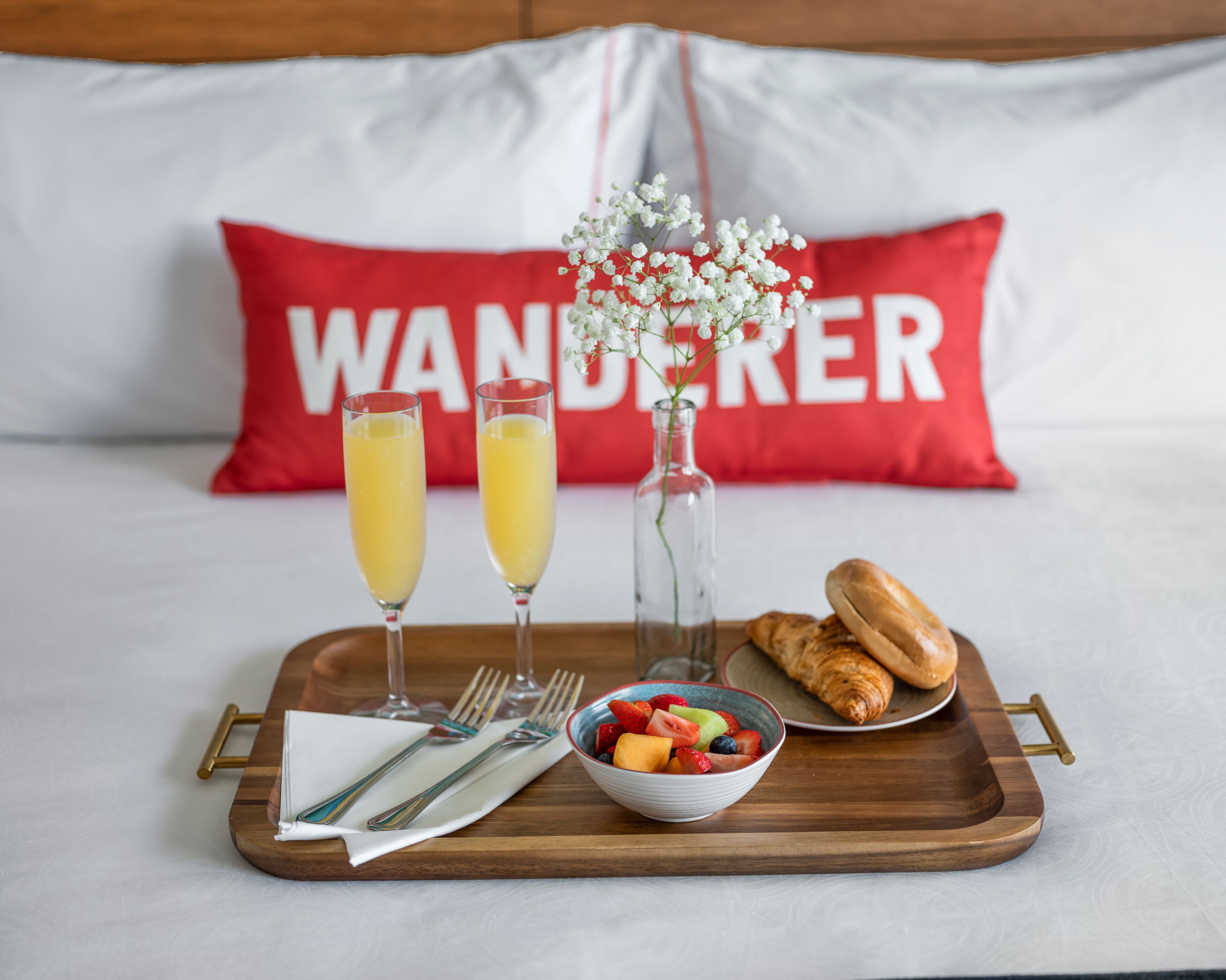 A breakfast tray with fruit, juice, a croissant, and a vase of flowers on a bed with a red pillow that says "WANDERER".