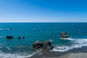 A coastal beach scene with clear blue water, rocky outcrops, and distant boats. - Ocean Front Enchanting Sunsets from  Wild Blue 2 (Brookings)