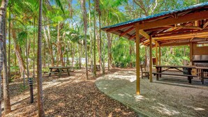 Un espace pique-nique abrité, doté de bancs et d’un barbecue, entouré de grands arbres et bordé d’un chemin de gravier.