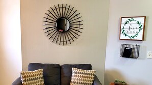 Ein dunkelbraunes Sofa mit gemusterten Kissen, ein flauschiger Teppich, ein runder Wandspiegel, ein kleiner Holzhochentisch und ein gerahmtes Schild an der Wand.