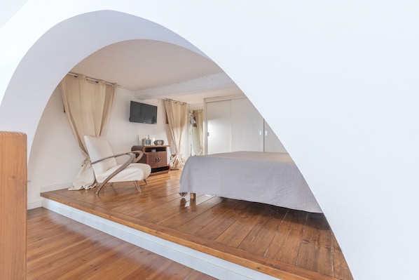 A bedroom with a bed, a chair, a desk, a TV, and a wooden floor.