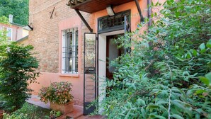 Un edificio de ladrillo con una pérgola de madera, una verja de metal y un jardín con plantas en macetas.