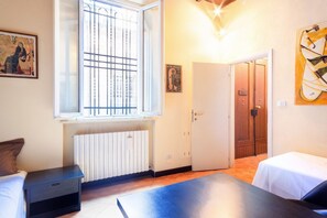 A room with a bed, a small table, a radiator, a window with bars, and a door leading to another room. - GuestHost - Ferrara Lilac Casa dell'Archivòlto with Garage (Ferrara)