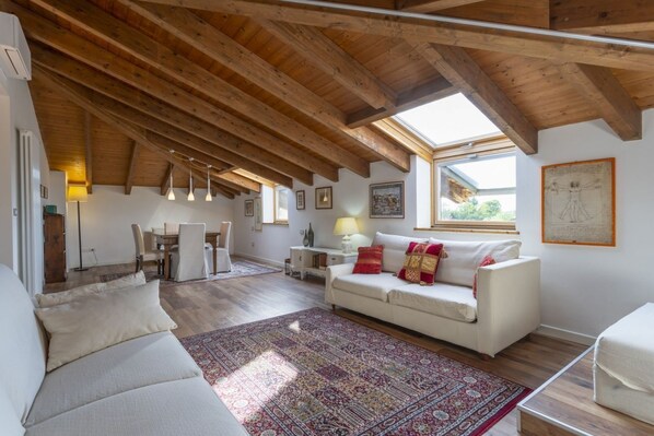 A spacious loft with a high ceiling, exposed wooden beams, a white sofa, a patterned rug, and a dining area with chairs and a table.
