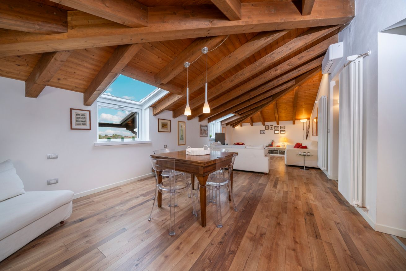 Un intérieur comprenant un coin repas, une cuisine et un espace de vie, avec des poutres en bois, une grande fenêtre et un canapé blanc.