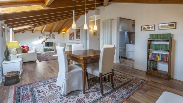 Un loft espacioso con techo alto, vigas de madera, un comedor con mesa y sillas, una cocina y una estantería.