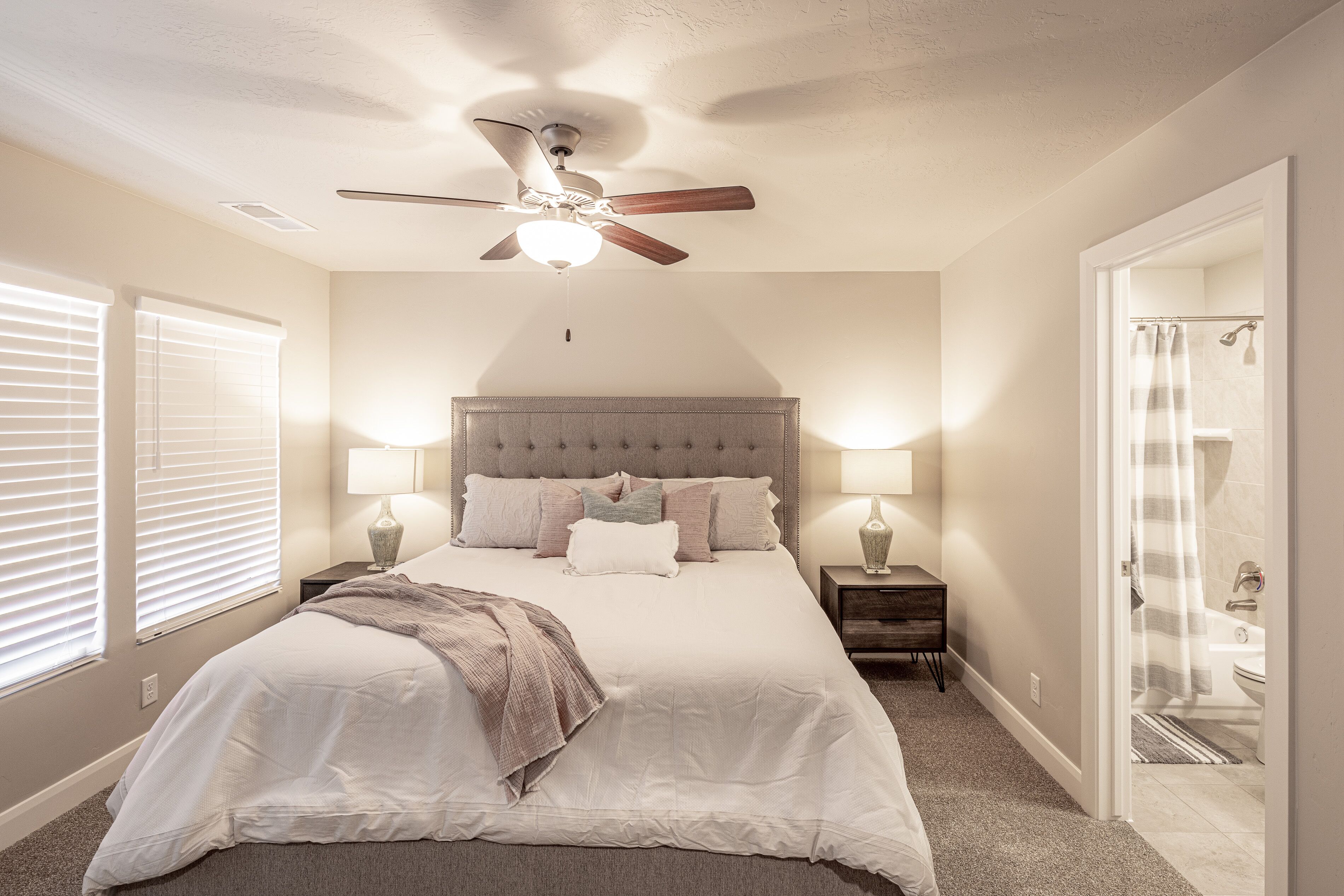 Une chambre à coucher avec un grand lit, deux tables de chevet avec des lampes, un ventilateur de plafond et une salle de bain visible à travers une porte ouverte.