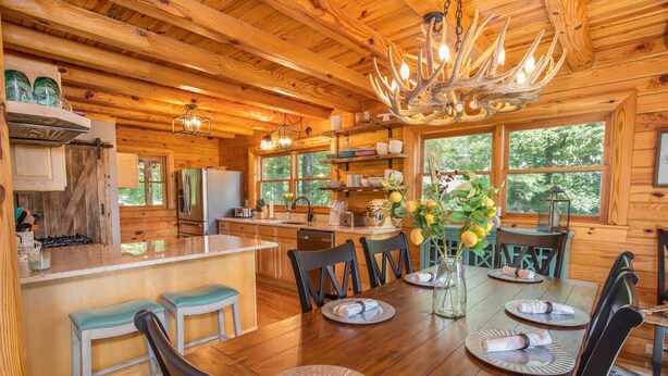 A rustic wooden cabin interior with a dining area, kitchen, and large windows. - Luxurious Log Cabin with Stunning Views (Sugar Mountain)