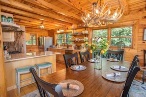 Cozy cabin-style interior with a warm aesthetic, featuring a central island and large windows offering views of the surrounding greenery.