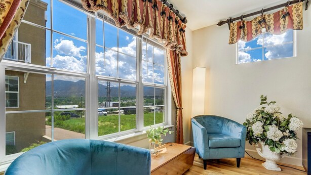 Habitación con un ventanal grande que ofrece vistas a montañas y vegetación. Hay un sillón azul y una silla más pequeña cerca del ventanal. Un banco de madera está situado frente al ventanal, y un jarrón con flores blancas reposa sobre él.