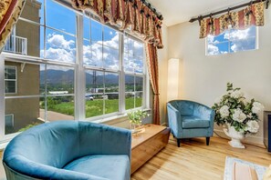 Habitación con un ventanal grande que ofrece vistas a montañas y vegetación. Hay un sillón azul y una silla más pequeña cerca del ventanal. Un banco de madera está situado frente al ventanal, y un jarrón con flores blancas reposa sobre él.