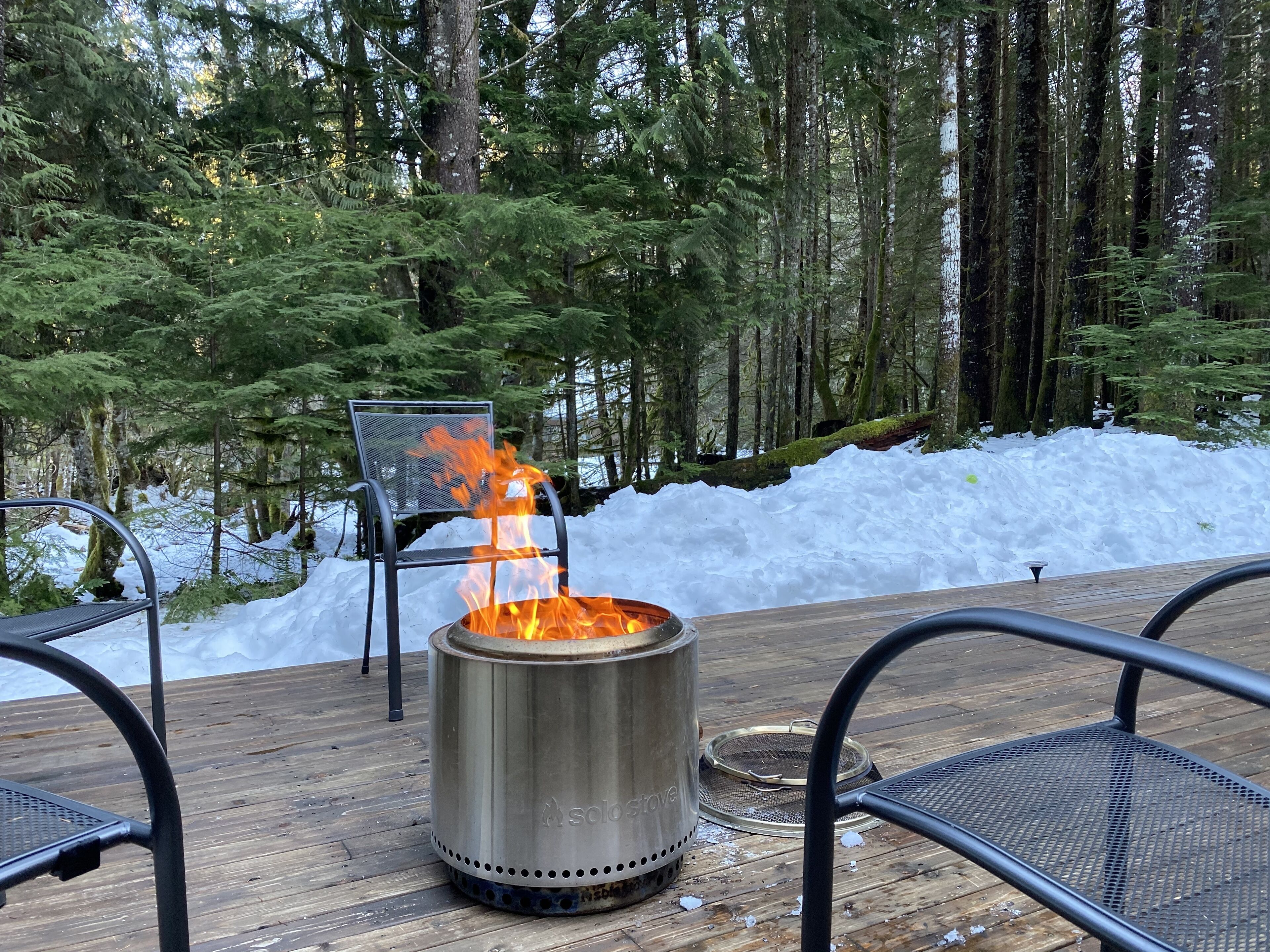 A metal fire pit with flames, surrounded by metal chairs on a wooden deck in a forested area with snow.