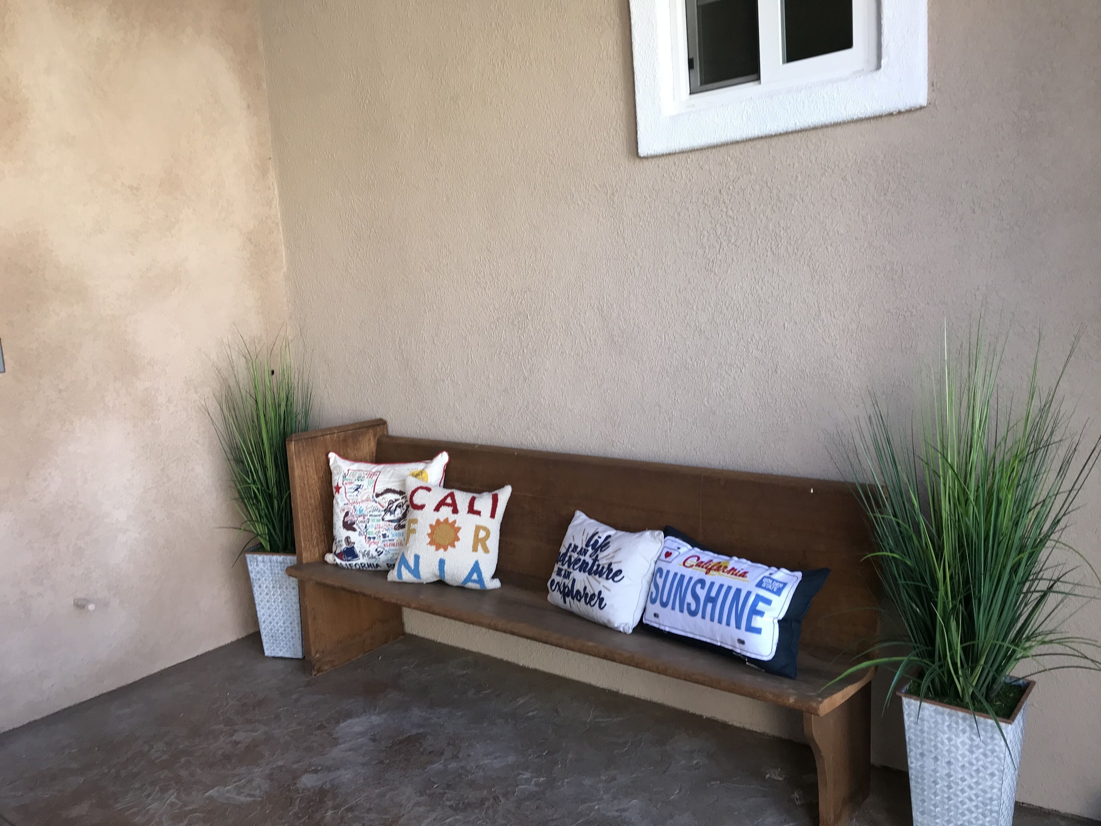 A wooden bench with two potted plants on either side, against a textured wall with a window above.
