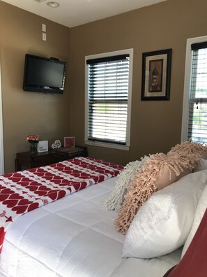 Un dormitorio con una cama, un televisor montado en la pared y dos ventanas con persianas.
