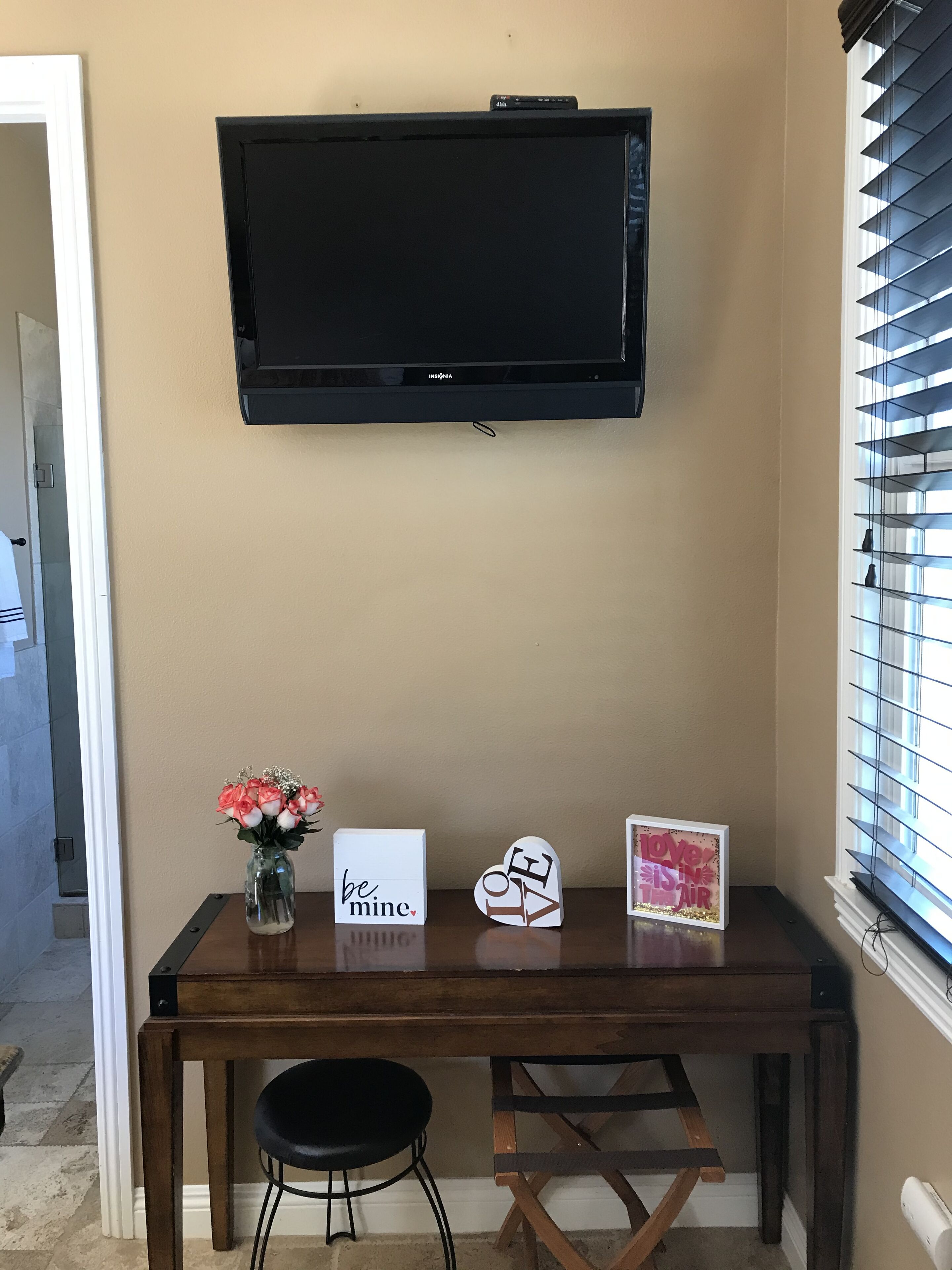 A room with a mounted flat-screen TV, a wooden desk with a stool, and decorative items including a vase of flowers and cards.