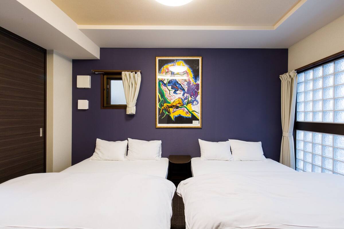 Ein Hotelzimmer mit zwei Betten, einem großen Fenster mit Vorhängen, einem Gemälde an der Wand und einer Deckenleuchte.