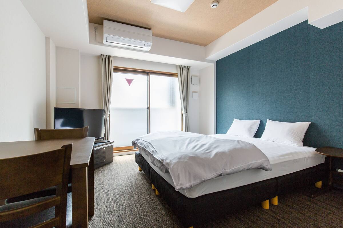 Ein Hotelzimmer mit Bett, Schreibtisch, Stuhl, Fernseher und einem Fenster mit Vorhängen.