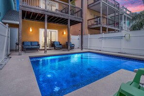 A swimming pool with a wooden deck and lounge chairs, surrounded by a white fence and a two-story building with balconies.