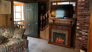 Una sala de estar con chimenea, un televisor montado encima, un sofá con estampado floral y una puerta que da a otra habitación.