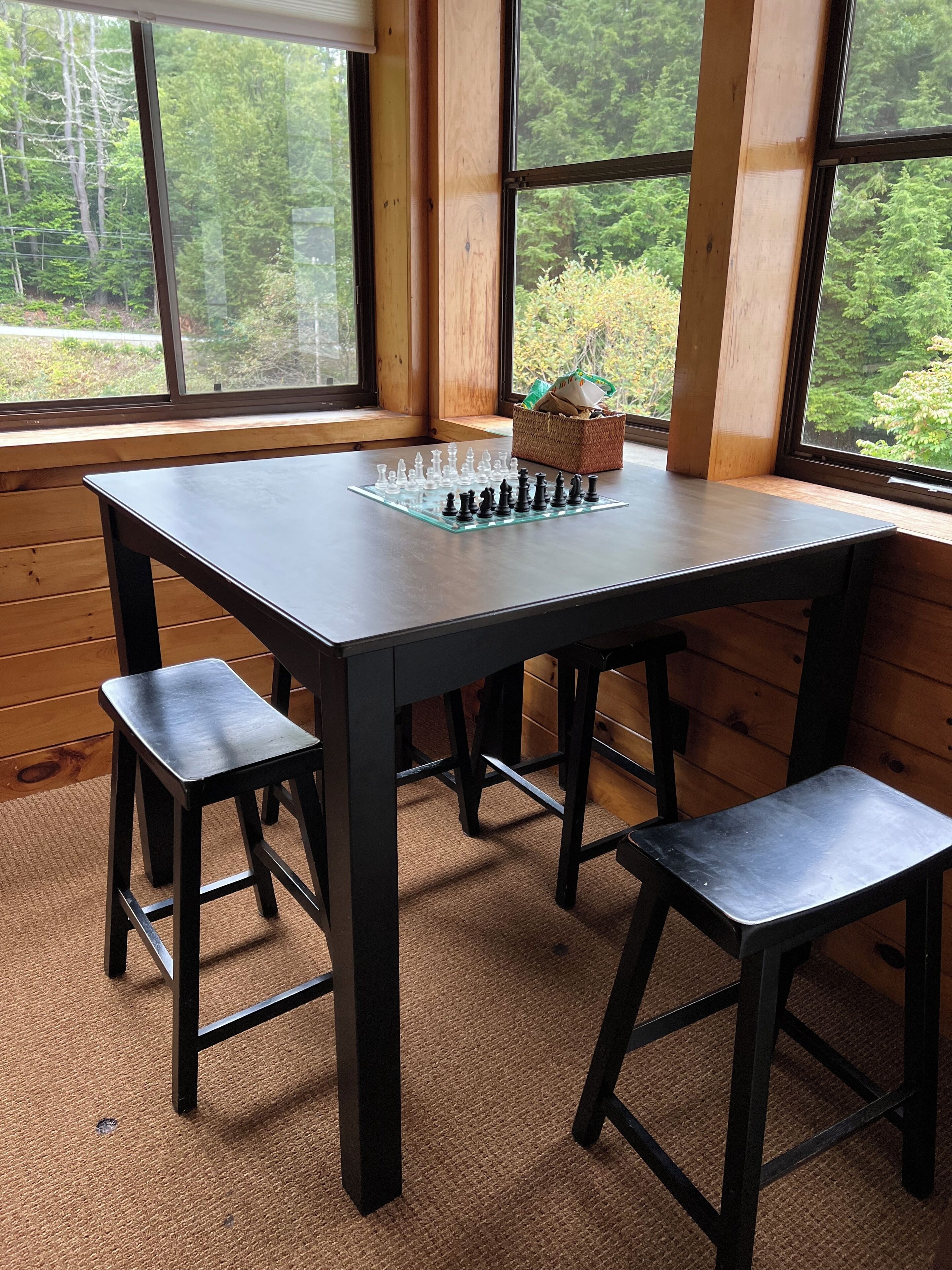 Une table en bois avec un jeu d’échecs, dans une pièce dotée de grandes fenêtres donnant sur un paysage arboré.