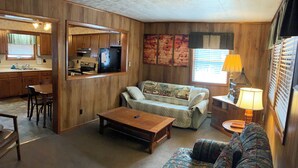 Una sala de estar acogedora con un sofá, una mesa de centro y un televisor. Hay una zona de comedor con una mesa y sillas, y una cocina con armarios y un refrigerador.