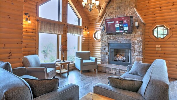 A log cabin interior with a stone fireplace, multiple seating areas, and a mounted TV. - Family Size Cabin with Kitchen, Fireplace, Hot Tub (Millersburg)