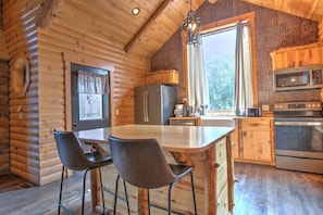 Une cabane en rondins dotée d’une cuisine comprenant un îlot central, des tabourets de bar, un réfrigérateur, un four à micro-ondes et un four traditionnel.