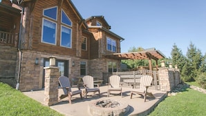 A two-story log cabin with a stone exterior, a covered patio with Adirondack chairs, and a fire pit. - Family Size Cabin with Kitchen, Fireplace, Hot Tub (Millersburg)