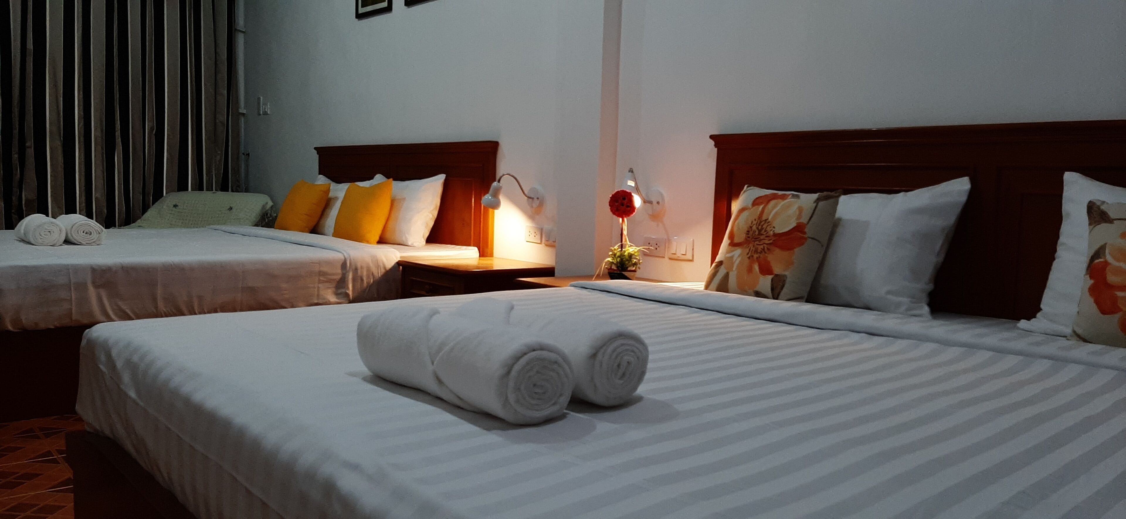 A hotel room with two beds, each with a wooden headboard, white linens, and rolled towels. There is a nightstand with a lamp and a small plant between the beds.