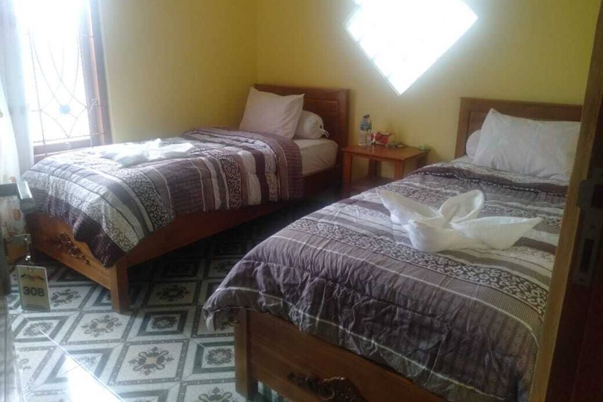 Two single beds with patterned bedding, a small bedside table with a bottle, and a window with a geometric view.