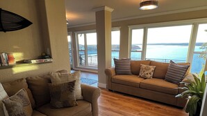 Una sala de estar con un sofá color beige, una mesa de centro y una estantería. Hay ventanas grandes con vistas al mar.