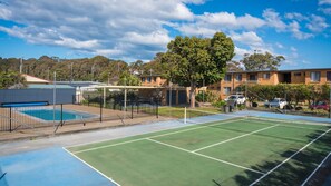 An outdoor tennis court with a pool, surrounded by a fence, and residential buildings in the background.
