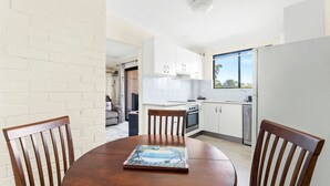 A modern kitchen with white cabinets, a stove, and a window. There is a dining table with chairs and a beach-themed placemat. - Apollo Unit 14 - 1st Floor (Narooma)