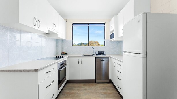 A modern kitchen with white cabinets, a stainless steel dishwasher, and a window with a view of the outdoors. - Apollo Unit 14 - 1st Floor (Narooma)