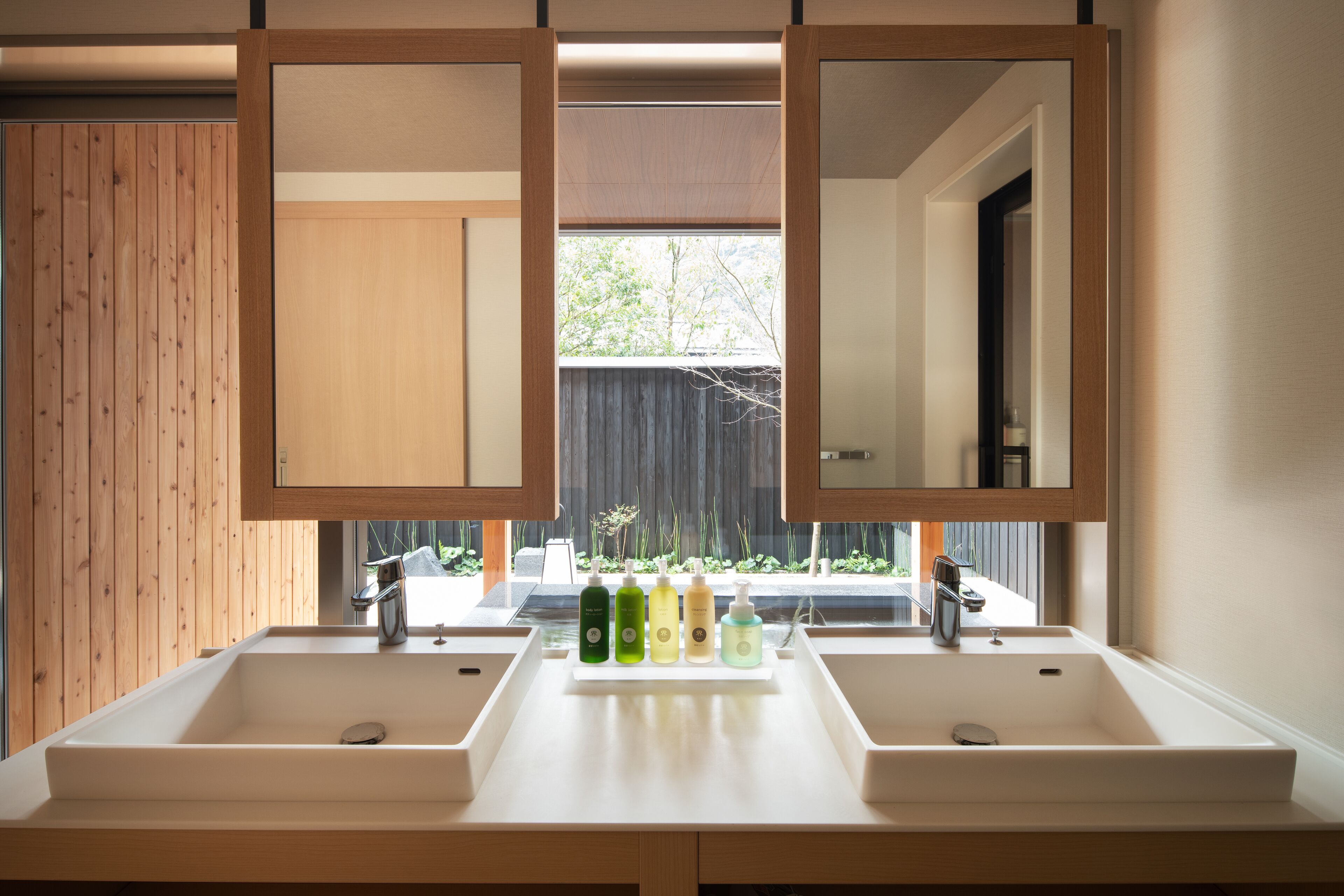 A bathroom with two sinks, a mirror, and a window with a view of greenery.