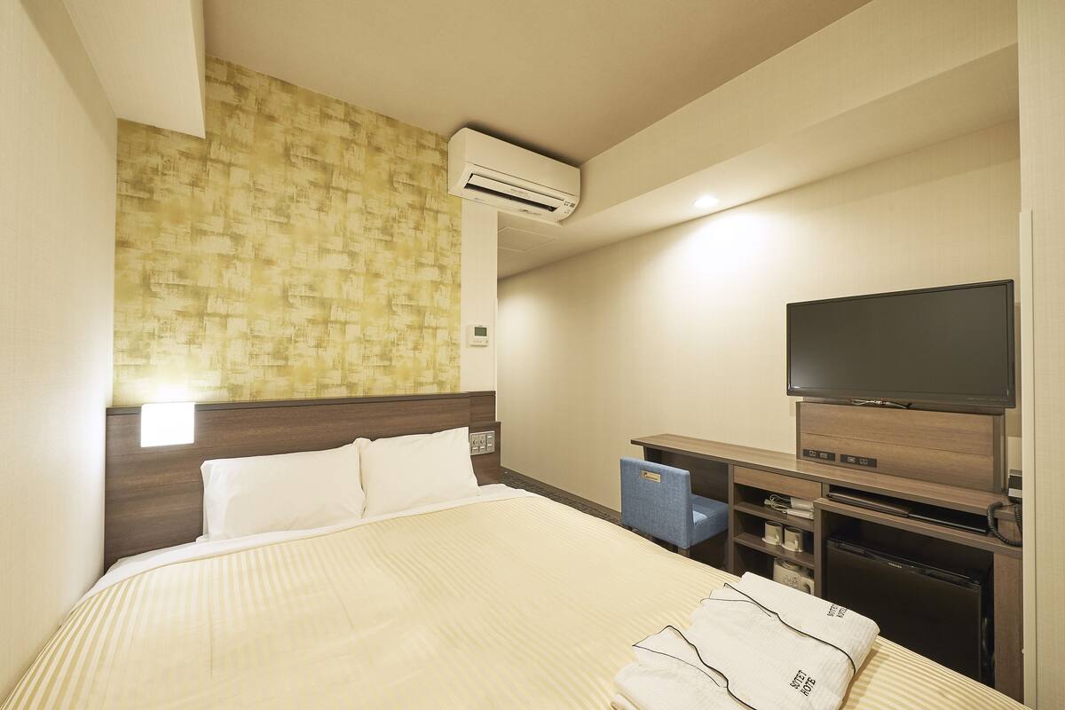A hotel room with a large bed, a flat-screen TV, a desk with a chair, and an air conditioning unit.